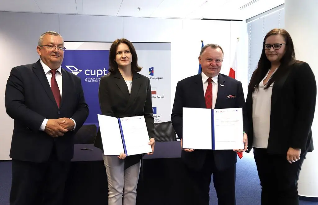 Cztery osoby (dwie kobiety i dwóch mężczyzn) stojące w rzędzie w sali konferencyjnej, trzymające podpisane dokumenty po uroczystym podpisaniu umowy w siedzibie CUPT.