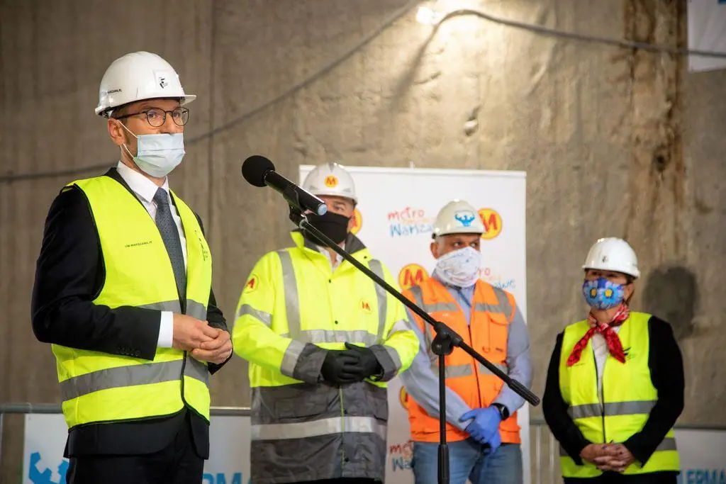 Przedstawiciel z delegacji uczestników przemawia do mikrofonu na placu budowy. Ma na sobie biały kask ochronny z warszawską syrenką, maseczkę na twarzy, marynarkę i żółtą kamizelkę odblaskową. W tle stoją trzy inne osoby, również w kaskach i kamizelkach roboczych. Tło stanowi surowa, betonowa ściana wykopu.