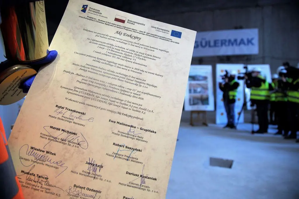 Zbliżenie na papierowy dokument Aktu Erekcyjnego z okazji budowy II linii metra w Warszawie. Widoczne są logotypy Funduszy Europejskich oraz podpisy oficjeli, w tym prezydenta Rafała Trzaskowskiego. W tle zamazane sylwetki uczestników uroczystości.