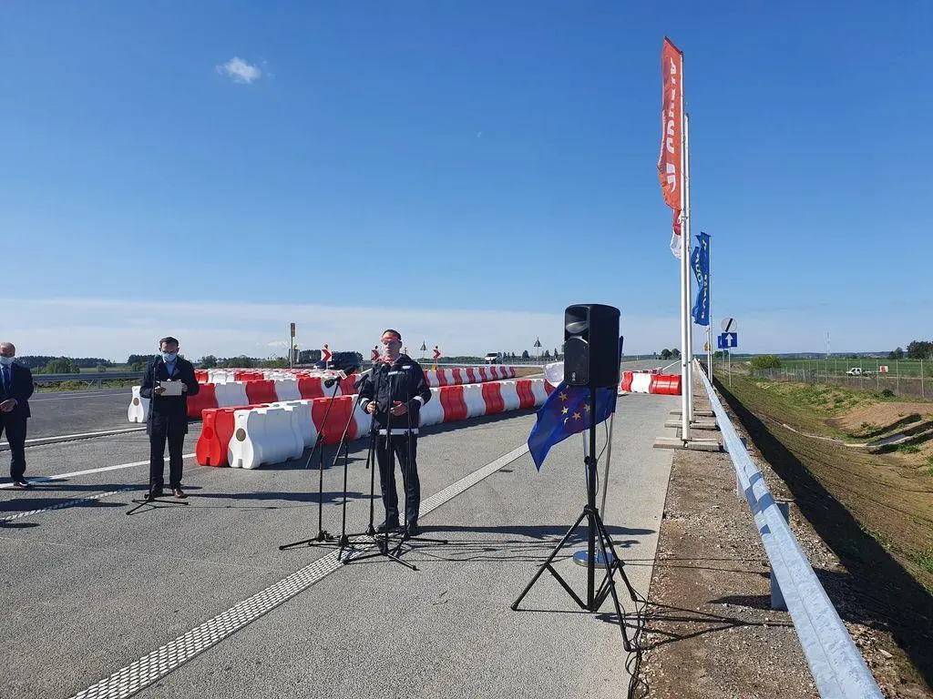 Konferencja na nowo wybudowanym odcinku drogi w słoneczny dzień. Mężczyzna w maseczce ochronnej przemawia do mikrofonów. Za nim, na asfalcie, ustawiono rzędy biało-czerwonych barier drogowych. Po prawej stronie stoi głośnik na statywie oraz powiewa flaga Unii Europejskiej. W tle widać inne osoby w maseczkach.