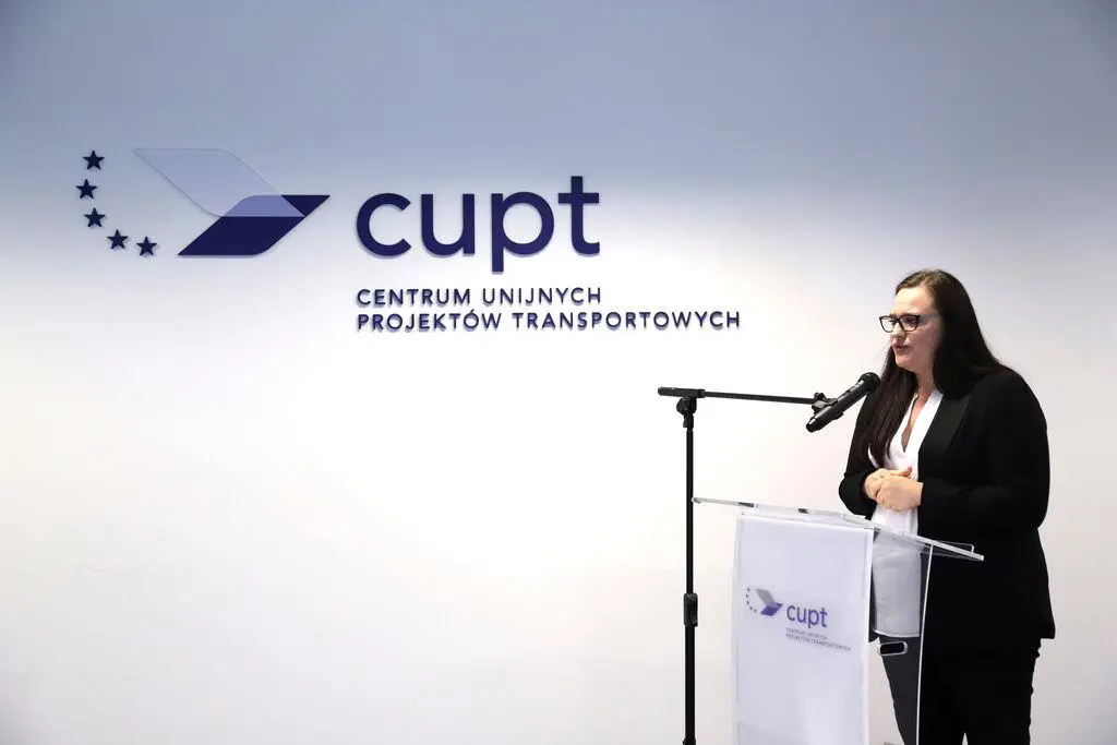 Kobieta w ciemnym żakiecie przemawiająca do mikrofonu podczas konferencji prasowej, stojąca za mównicą z logo CUPT na tle białej ściany z napisem „Centrum Unijnych Projektów Transportowych