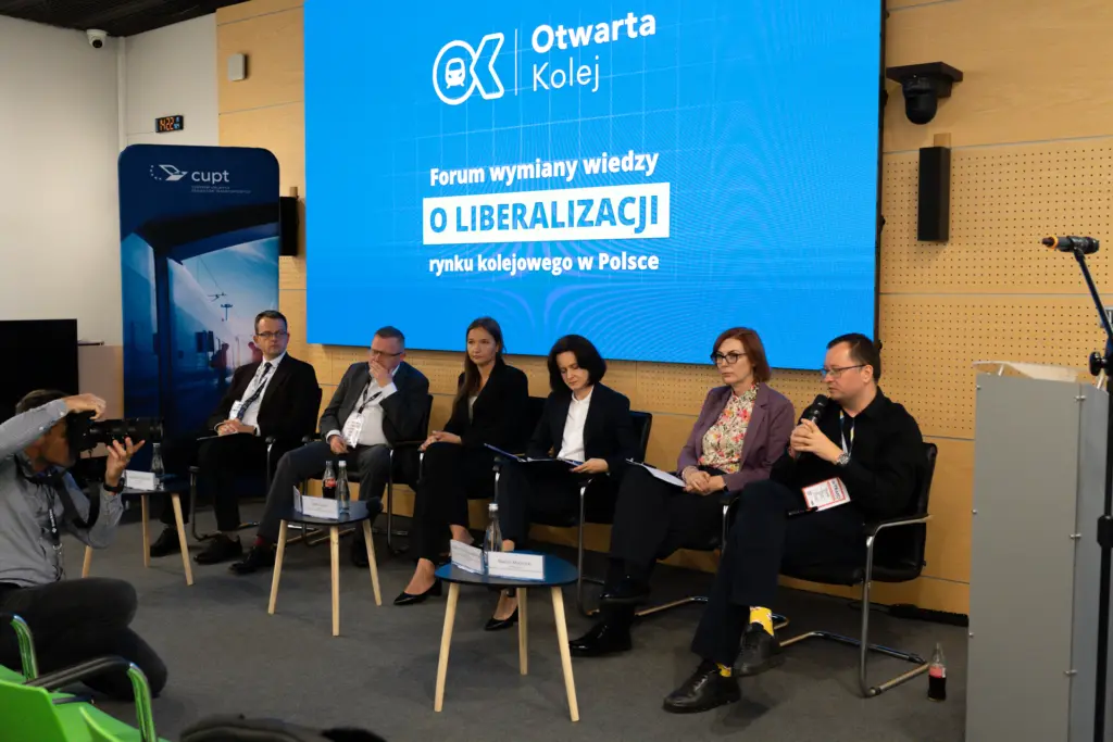 Panel dyskusyjny na scenie. Sześcioro prelegentów siedzi obok siebie, a mężczyzna po prawej stronie wypowiada się przez mikrofon. W tle widoczny jest duży, niebieski ekran z logo „Otwarta Kolej” oraz napisem: „Forum wymiany wiedzy O LIBERALIZACJI rynku kolejowego w Polsce”. Po lewej stronie znajduje się pionowy baner z logo CUPT, a przed sceną fotograf robi zdjęcia uczestnikom panelu.